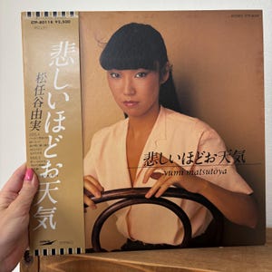 以下が含まれることがあります： 女性が写った、薄いピンク色のトップスを着たヴィンテージレコードアルバムのカバー。アルバムタイトル「悲しいほどお天気」とアーティスト名「松任谷由実」が日本語で表示されています。レコードは段ボールのスリーブに入っています。