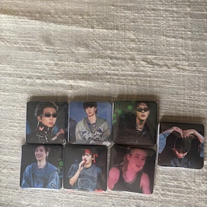 May include: A collection of seven square-shaped photo cards featuring images of a musical group. Each card displays a different member in various poses and outfits. The cards are arranged on a neutral-colored fabric surface.