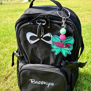Può includere: Zaino nero con logo "Infinity" bianco e testo "Brooklyn". Un portachiavi a forma di fiocco verde e rosa con il nome "Pebbles" è appeso alla cerniera. Il portachiavi ha una perla rosa e una verde.