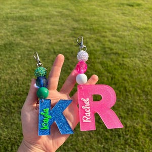 May include: Two glittery keychains, one blue with a green "Kayla" and a "K", the other pink with a white "Rachel" and a "R". Each keychain has a beaded accent and a silver clasp.
