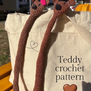 Könnte beinhalten: Zwei braune Häkelteddybären mit langen Hälsen, einer mit einer rosa Schleife, der andere mit einem schwarzen Auge. Die Bären stehen vor einer beigefarbenen Tragetasche. Auf der Tasche steht "Teddy crochet pattern" mit einem Herzsymbol.