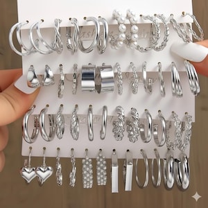 Può includere: Una collezione di orecchini argentati esposti su una scheda bianca. Gli orecchini includono vari stili come cerchi, design intrecciati e quelli adornati con perle finte. Il set offre una varietà di dimensioni e forme.