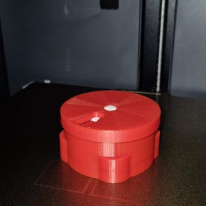 May include: A 3D-printed red plastic object with a circular top and a rectangular base. The top has a white circular insert. The object is sitting on a black surface.