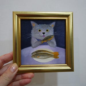 May include: A framed painting depicts a gray cat holding a fish, with two more fish on a plate below. The cat has yellow eyes and a blue collar. The painting is in a gold frame, creating a whimsical and artistic piece.