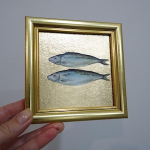 Pode incluir: Uma obra de arte quadrada emoldurada apresentando dois peixes em tons de azul pintados sobre um fundo de folha de ouro texturizado. A moldura é de cor dourada polida com um perfil simples e limpo, envolvendo a ilustração central dos peixes.