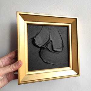 May include: An abstract artwork in a gold frame. The piece features a textured black design on a black background. The artwork has a modern aesthetic and is suitable for home decor, with a focus on texture and form.