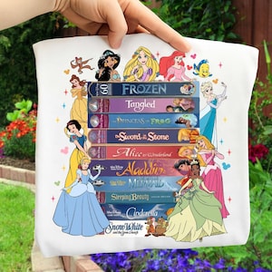Puede incluir: Un bolso tote blanco con ilustraciones de princesas de Disney y cintas VHS de películas clásicas de Disney. Los títulos de las cintas incluyen "Frozen", "Tangled" y "Aladdin". El bolso es sostenido por una mano.