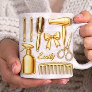 May include: White ceramic mug featuring gold-colored beauty tools: hair dryer, scissors, comb, spray bottle, and hair clips. A gold bow and the name "Emily" are also displayed. The mug has a pearlescent background.