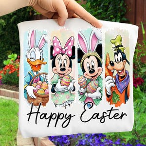 May include: White tote bag featuring Donald Duck, Minnie Mouse, Mickey Mouse, and Goofy dressed as Easter bunnies. Each character holds an Easter egg. The words "Happy Easter" are written in black script across the bottom.