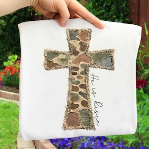May include: White fabric with a cross design. The cross is made of patchwork fabric in shades of brown, green, and tan. The text "He is Risen" is written in black script on the fabric. The item is held up by a hand.
