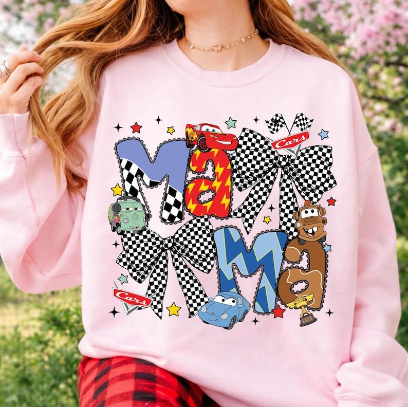 May include: Pink sweatshirt with the word "Mama" in a Cars-themed design. The letters are decorated with checkered patterns, lightning bolts, and images of cartoon cars. The design includes stars and checkered flag accents.