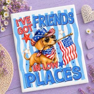 Könnte beinhalten: Ein rechteckiger Druck mit einem hellbraunen Dackel, der einen sternenbesetzten Cowboyhut, eine Sonnenbrille und ein passendes Halstuch trägt. Der Hund hält eine amerikanische Flagge. Der Text lautet I'VE GOT FRIENDS IN LOW PLACES auf blau-weiß gestreiftem Grund.