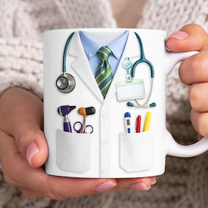 Könnte beinhalten: Weiße Keramik-Tasse mit einem Design eines Arztkittels. Das Design umfasst ein blaues Hemd, eine grün gestreifte Krawatte, ein Stethoskop und Taschen mit medizinischen Werkzeugen.
