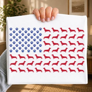 Op de afbeelding: Een wit rechthoekig stoffen item met een grafisch patroon dat is gerangschikt als de Amerikaanse vlag. Het kanton bestaat uit blauwe pootafdrukken, terwijl de strepen zijn samengesteld uit rijen rode teckelsilhouetten met een textuur.
