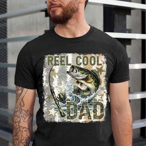 May include: Dark gray t-shirt featuring a fishing-themed graphic. The design includes a leaping fish, fishing rod, and the text "REEL COOL DAD" in a camouflage style font. The graphic is set against a distressed background with star accents.