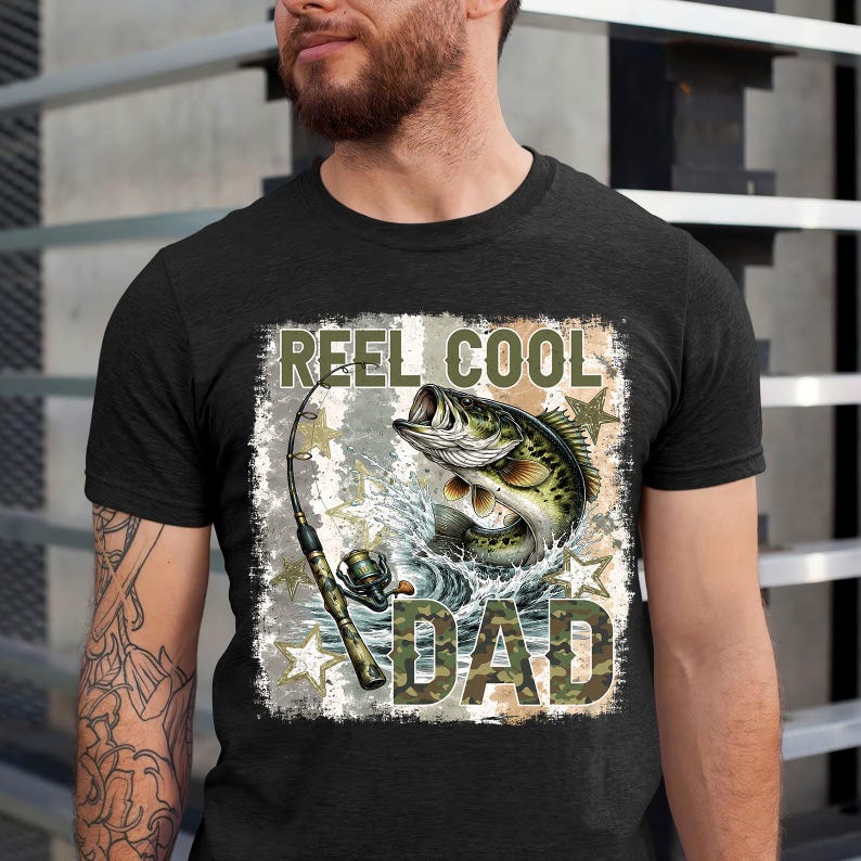 May include: Dark gray t-shirt featuring a fishing-themed graphic. The design includes a leaping fish, fishing rod, and the text "REEL COOL DAD" in a camouflage style font. The graphic is set against a distressed background with star accents.