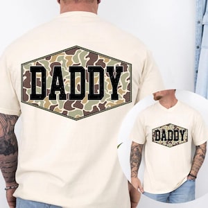 May include: A cream-coloured t-shirt with a camouflage pattern inside a black-outlined hexagon. The word "DADDY" is printed in bold, black letters. The shirt has short sleeves and a crew neck.