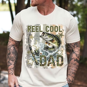 May include: Cream-colored t-shirt featuring a graphic of a largemouth bass leaping from water, with a fishing rod. The text "REEL COOL DAD" is printed above and below the fish in a camouflage font, with star accents.