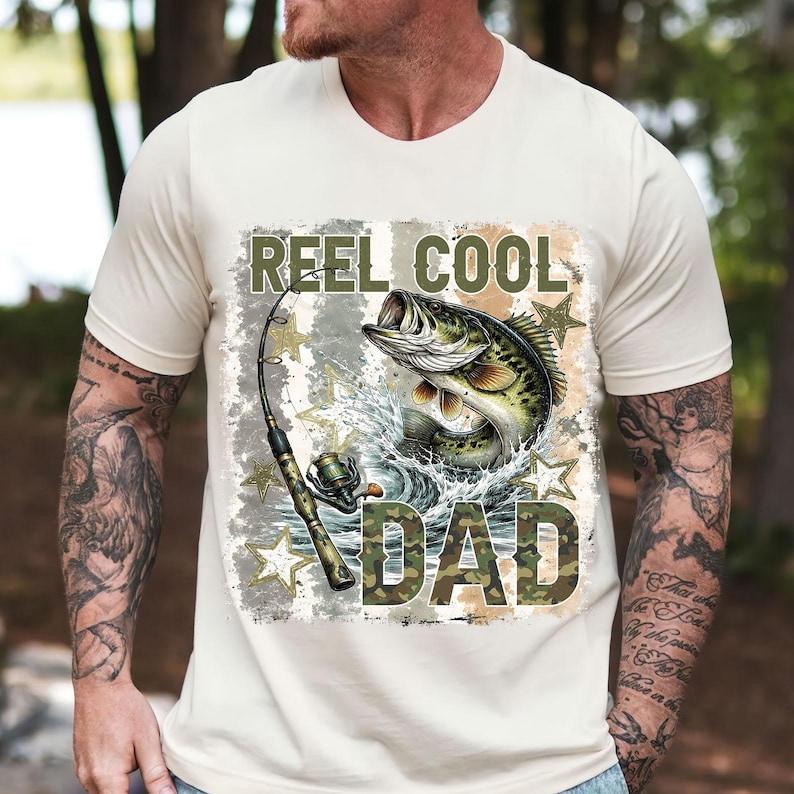 May include: Cream-colored t-shirt featuring a graphic of a largemouth bass leaping from water, with a fishing rod. The text "REEL COOL DAD" is printed above and below the fish in a camouflage font, with star accents.