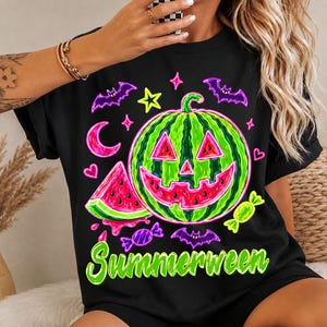 Op de afbeelding: Een zwart t-shirt met een neon-stijl afbeelding van een watermeloen uitgesneden als een pompoen, een watermeloenpartje, paarse vleermuizen, een roze maansikkel, gele sterren, snoepjes en de groene tekst Summerween onderaan gedrukt.