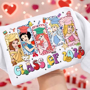 Puede incluir: Sudadera blanca con un gráfico colorido de princesas de Disney con gorros de graduación. El diseño incluye el texto "CLASS OF 2026" en una fuente lúdica, con varios elementos decorativos.