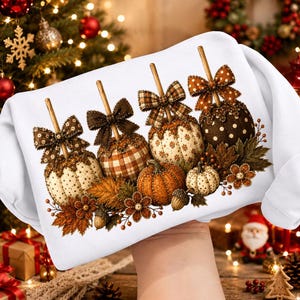May include: A white sweatshirt featuring a graphic print of four pumpkins on sticks, each decorated with a patterned bow. The pumpkins display various textures including polka dots and plaid, surrounded by autumn leaves, acorns, and berries.