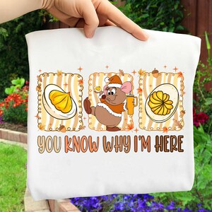 May include: White t-shirt with a cartoon mouse in a Santa hat, holding a mug, and two egg halves. The text "YOU KNOW WHY I'M HERE" is printed in a brown and orange font. The design has a fall theme.