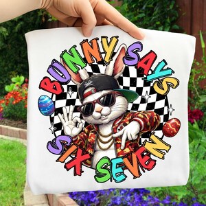 May include: White tote bag featuring a cartoon rabbit wearing sunglasses, a backwards cap, and a camouflage jacket. The text "Bunny Says Six Seven" is in a colorful, circular design with a checkered background and Easter eggs.
