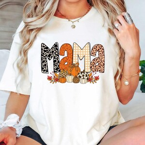 Puede incluir: Camiseta color crema con la palabra "MAMA" en un dise&ntilde;o de tem&aacute;tica oto&ntilde;al. Las letras tienen estampados de animales, naranja y cuadros. Calabazas y follaje de oto&ntilde;o est&aacute;n debajo de la palabra, creando un gr&aacute;fico estacional.