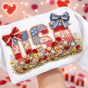May include: White pillow with the word "USA" appliqued in patriotic patchwork fabric. The letters are adorned with bows and surrounded by a floral border of red poppies, daisies, and other flowers. The background features blurred heart shapes.