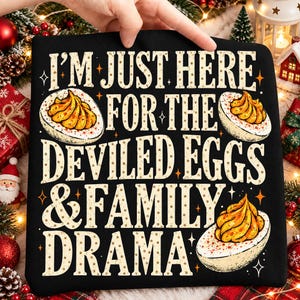 May include: A black sweatshirt featuring the text I'M JUST HERE FOR THE DEVILED EGGS & FAMILY DRAMA in a beige, dotted font. The design includes three illustrations of halved deviled eggs with yellow yolks and orange star accents.