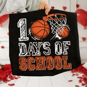 May include: Black t-shirt with a basketball theme. The design includes the text "100 DAYS OF SCHOOL" in white and orange, along with basketballs and a hoop.