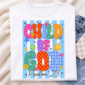 May include: White t-shirt with the words "CHILD OF GOD" in colorful, patterned letters. The design includes stars, flowers, and the text "Psalm 23" on a light blue striped background.
