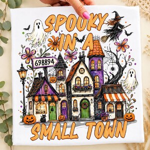 May include: A white t-shirt featuring a colorful Halloween graphic with the text "SPOOKY IN A SMALL TOWN" and "698894". The design includes a haunted village, ghosts, a witch on a broomstick, jack-o'-lanterns, a black cat, and purple flowers.