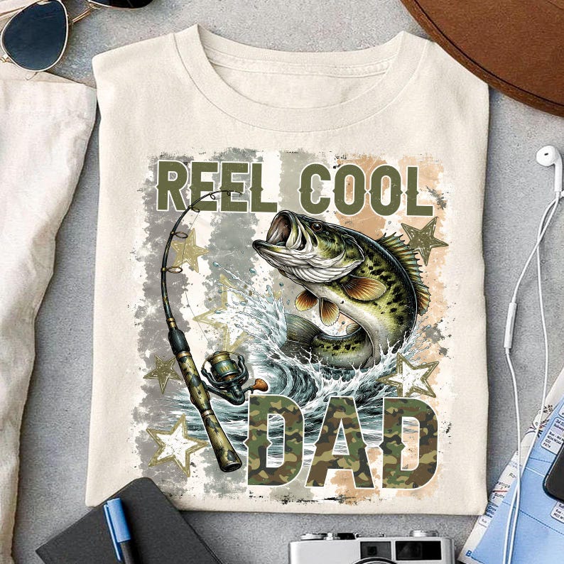 May include: Cream-colored t-shirt with a graphic featuring a largemouth bass leaping from water, a fishing rod, and the text "REEL COOL DAD" in camouflage lettering. The design includes stars and water splashes.