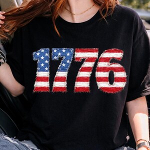 May include: Black t-shirt featuring the number "1776" in a patriotic design. The numbers are styled with the American flag, using red and white stripes and blue with white stars. The shirt is a casual style.