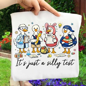May include: White tote bag featuring a cartoon illustration of ducks dressed as a scientist, student, and teacher. The text "It's just a silly test" is written in black script. The ducks are holding school supplies.