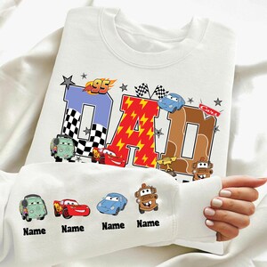Peut inclure: Sweat-shirt blanc avec un motif "DAD" coloré représentant des voitures de course et des éclairs. Le motif comprend le chiffre "95" et des drapeaux à damier. La manche affiche quatre voitures de dessin animé avec des noms personnalisés.