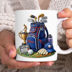 May include: White ceramic mug featuring a golf-themed design. The design includes a blue and red golf bag, golf clubs, a trophy, a golf ball, a score calculator, a blue cap, and golf gloves. The mug is held by a person.