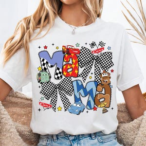 May include: White t-shirt with the word "MAMA" in colorful, cartoon-style lettering. The letters are decorated with race cars, checkered bows, and stars. The word "Cars" is also printed on the shirt.