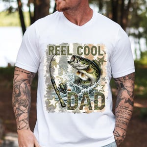 May include: White t-shirt with a graphic featuring a leaping fish, fishing rod, and the words "REEL COOL DAD" in camouflage lettering. The design includes water splashes and star accents, perfect for fishing enthusiasts.