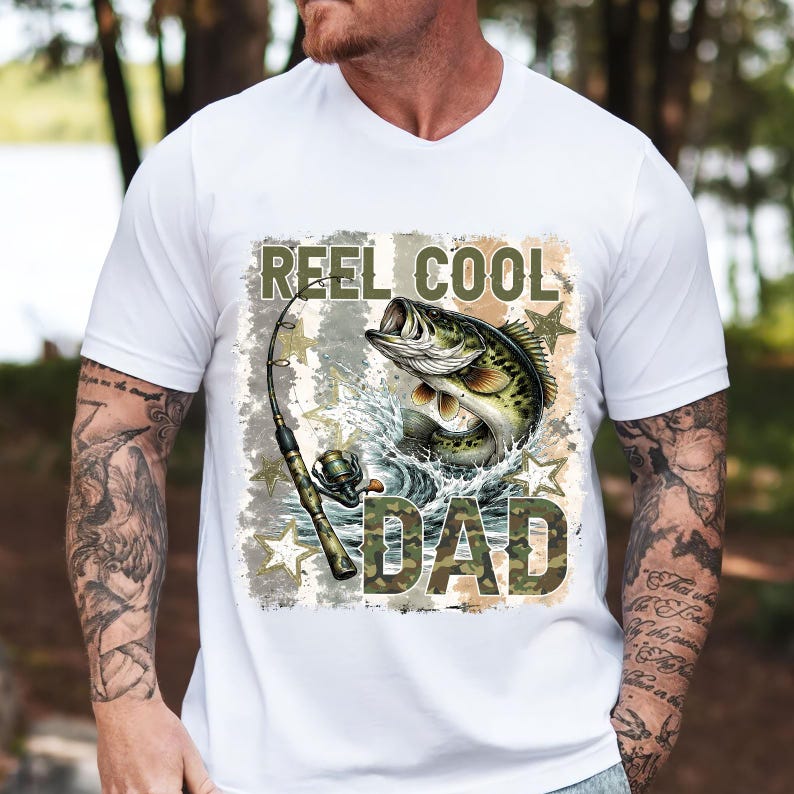 May include: White t-shirt with a graphic featuring a leaping fish, fishing rod, and the words "REEL COOL DAD" in camouflage lettering. The design includes water splashes and star accents, perfect for fishing enthusiasts.