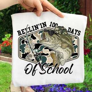 May include: White tote bag featuring a graphic of a bass wearing sunglasses and a camouflage hat. The design includes a fishing rod and the text "REELIN' IN 100 DAYS OF SCHOOL" in black script. The background has a camouflage pattern.