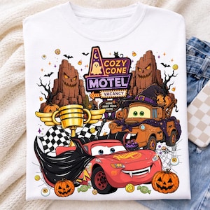 May include: A white t-shirt featuring a graphic print of a red race car wearing a black cape and a brown tow truck wearing a witch hat. The design includes a Cozy Cone Motel sign, jack-o'-lanterns, a gold trophy, and checkered flags.