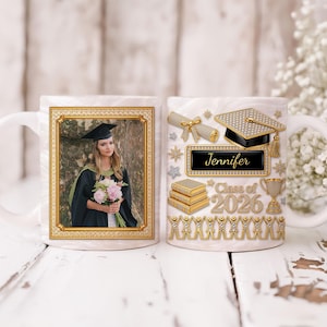 Op de afbeelding: Witte keramische afstudeerbeker met een foto van een afgestudeerde in toga en hoed, goudkleurig omlijst. De andere kant heeft gouden afstudeer-thema versieringen, waaronder een mortelbord en de tekst "Jennifer Class of 2026."