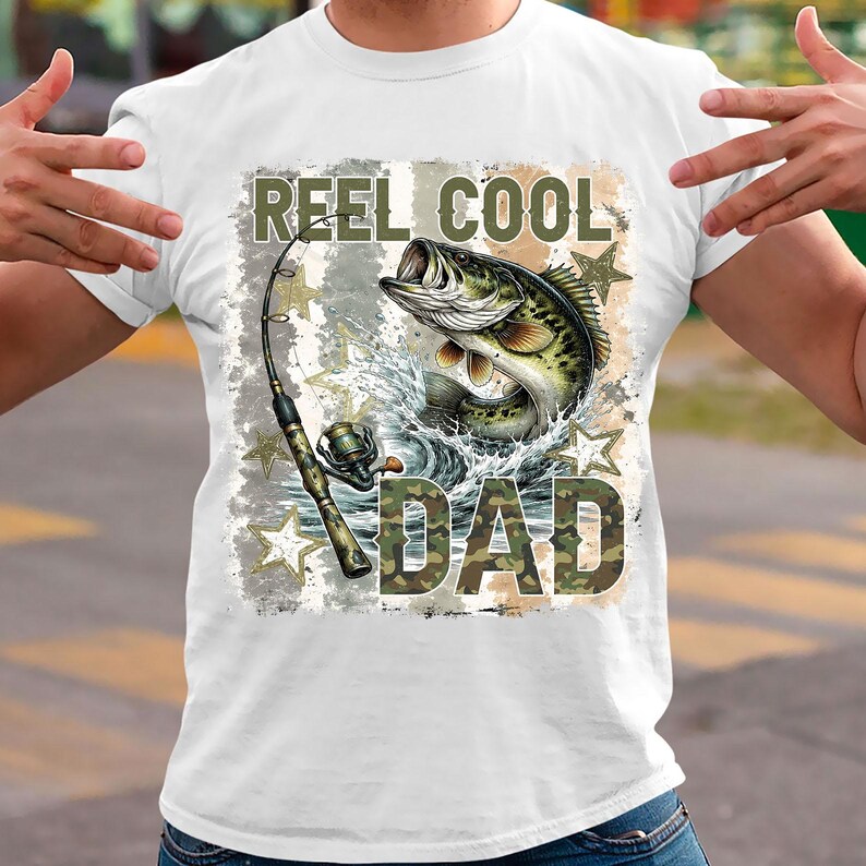 May include: White t-shirt with a fishing-themed graphic. The design features a leaping fish, fishing rod, and the words "REEL COOL DAD" in camouflage lettering. The graphic includes water splashes and star accents.