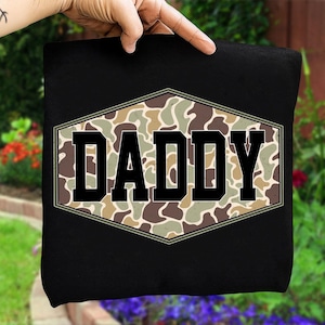 May include: Black t-shirt featuring the word "DADDY" in bold, black letters within a camouflage design. The camouflage is in a hexagonal shape. The background shows a garden with flowers.