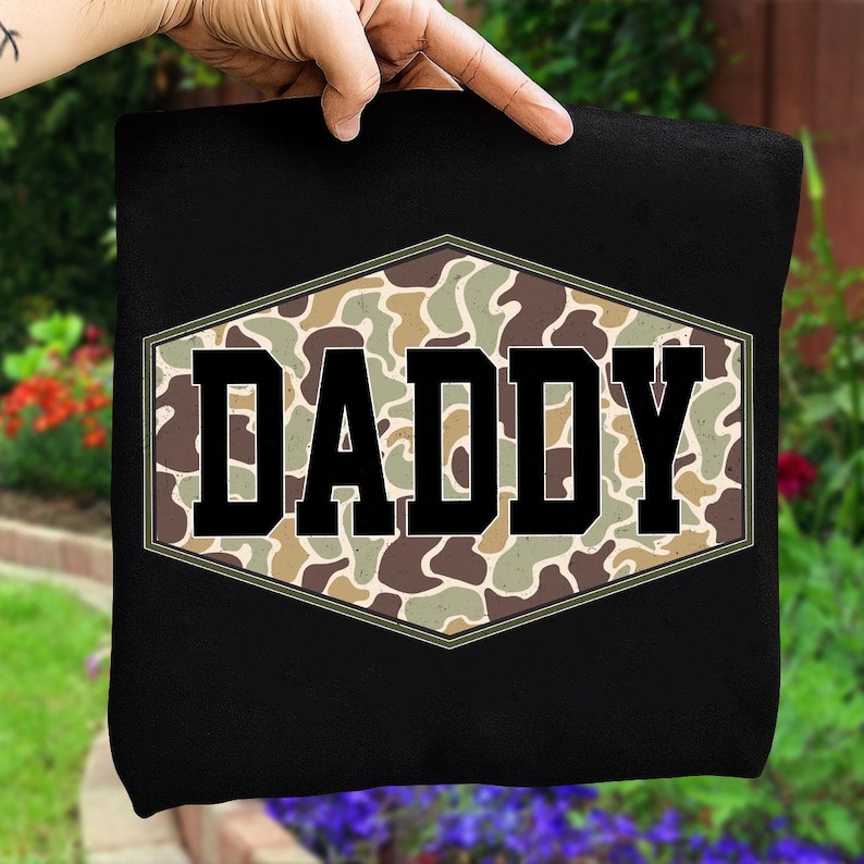 May include: Black t-shirt featuring the word "DADDY" in bold, black letters within a camouflage design. The camouflage is in a hexagonal shape. The background shows a garden with flowers.