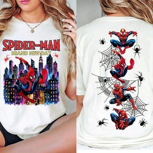 May include: A white t-shirt featuring a graphic print of Spider-Man on the front and back. The front displays the text "SPIDER-MAN BRAND NEW DAY" above Spider-Man leaping in front of a city skyline. The back shows four Spider-Man figures and spiders.