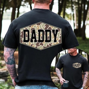 May include: Black t-shirt with a camouflage design in a hexagonal shape on the back, featuring the word "DADDY" in bold, black letters. The shirt is short-sleeved and appears to be made of cotton.